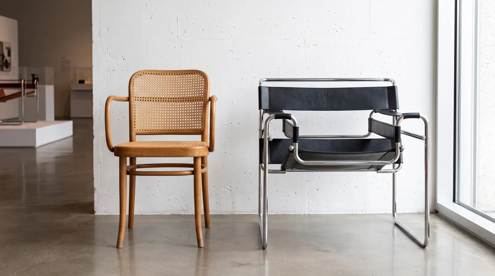 Chaise en bois courbé et cannage à côté d'un fauteuil Wassily en tubes d'acier chromé et sangles de cuir noir, sur sol de béton ciré devant un mur blanc